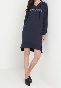 Granatowa sukienka z kapturem i dekoltem w kształcie litery V, ozdobiona logo na piersiach, z bocznymi rozcięciami i długimi rękawami. Wykonana z miękkiego, fakturowanego materiału.