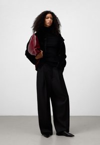 Zwarte, gestructureerde, oversized jas met knopen, zwarte coltrui, brede zwarte broek en een bordeauxrood tasje. Wijde, zwarte schoenen maken de look compleet.