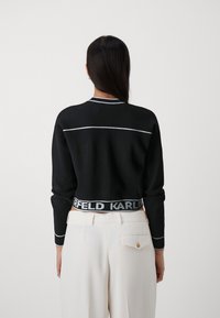 Maglione nero con cuciture a contrasto bianche, polsini a coste e design cropped. Presenta una vita con logo in nero e grigio.
