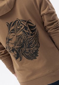 Felpa con cappuccio in tessuto marrone chiaro, caratterizzata da una grande grafica di un leone nero con dettagli intricati e modelli di foglie sulla parte posteriore.