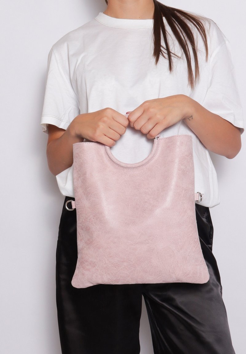 Blush roze, getextureerde neplederen tote bag met een afgeronde handvat en een hoofd ritsluiting, vastgehouden door een persoon die een wit t-shirt draagt.