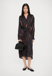Vestido longo preto com padrões coloridos em forma de onda, ajustado na cintura com um cinto correspondente. Usado com sapatilhas pretas e uma pequena bolsa.