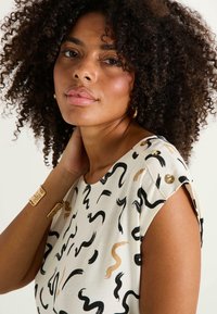 Blusa blanca con patrones en forma de garabato en negro y dorado. Presenta botones dorados en el hombro. La modelo tiene el cabello rizado y lleva una pulsera dorada.