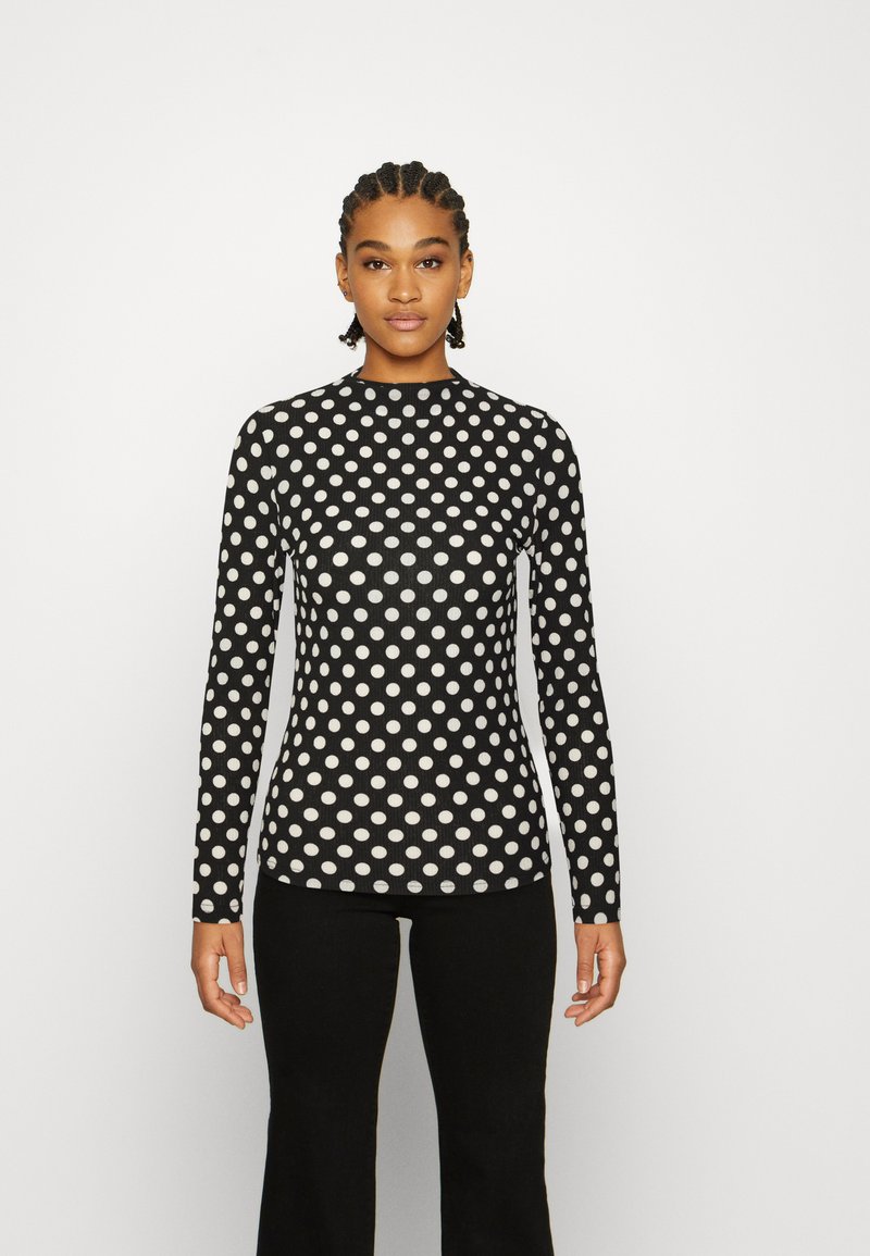 Blusa de mangas compridas ajustada em preto com bolinhas brancas, textura canelada e decote alto. Combinada com calças flare pretas.