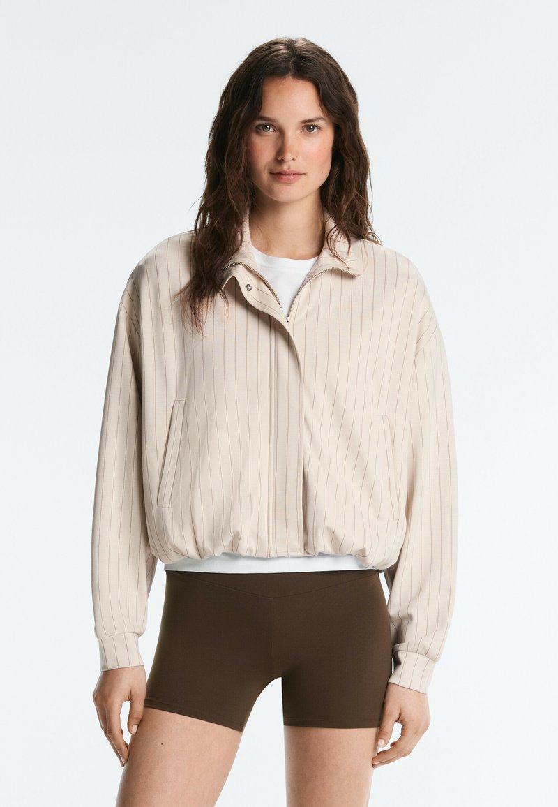 Femme aux longs cheveux bruns portant une veste zippée beige à fines rayures sur une chemise blanche et un short marron foncé ajusté, debout devant un fond uni.