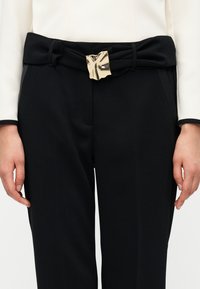Μαύρο παντελόνι με φαρδιά μπατζάκια και ζώνη με κόμπο, διακοσμημένη με λαμπερή, ακανόνιστη χρυσή αγκράφα. Λείο ύφασμα με εφαρμοστή γραμμή.