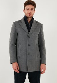 Cappotto di lana grigio con due tasche anteriori, tre bottoni e rever a punta. Indossato sopra un capo interno con zip di colore blu navy per maggiore calore.