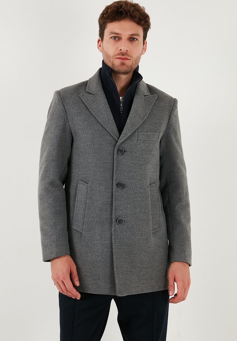 Cappotto di lana grigio con due tasche anteriori, tre bottoni e rever a punta. Indossato sopra un capo interno con zip di colore blu navy per maggiore calore.