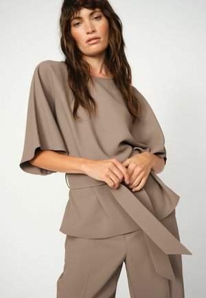 Blusa taupe con maniche larghe e una cintura in vita, caratterizzata da una texture liscia. Abbinata a pantaloni coordinati in un colore simile.