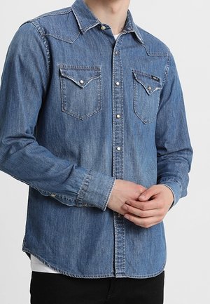 Homme portant une chemise en jean boutonnée avec deux poches poitrine, debout les mains doucement jointes devant lui, sur un fond uni.