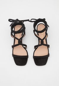 Sandales à brides en daim noir avec bout carré, liens à la cheville et semelles intérieures beiges avec marquage texte doré.