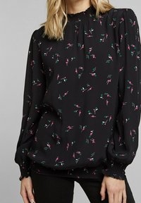 Schwarze Bluse mit langen, gepufften Ärmeln und einem gerafften Ausschnitt. Verfügt über kleine pinke und grüne Blumenmuster überall. Weicher Stoff.