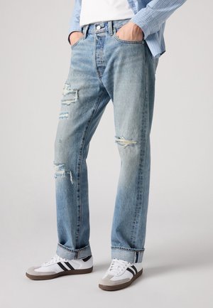 Man met lichtblauwe gescheurde spijkerbroek met opgerolde pijpen en witte sneakers met zwarte strepen, handen in de zakken.