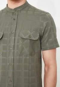 Lyhythihainen kauluspaita oliivinvihreänä, jossa on ruutuprintti. Paidassa on kaksi rintataskua ja nappidetaljit. Kangas on sileä ja kevyt.