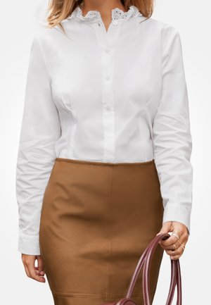 Chemise blanche à manches longues avec boutons et détails brodés sur le col, associée à une jupe en cuir marron ajustée. La personne tient un sac à main bordeaux.