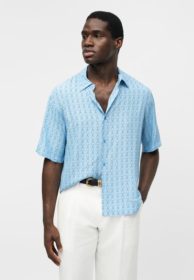 Άνδρας που φοράει ανοιχτό μπλε πουκάμισο με κοντό μανίκι και κυκλικό σχέδιο, λευκό παντελόνι και μαύρη ζώνη, κοιτάζει προς τα αριστερά του.