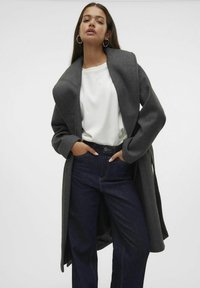 Abrigo gris de gran tamaño con un cuello ancho, llevado sobre un suéter blanco y unos vaqueros de tiro alto azul oscuro, con mangas largas y un ajuste relajado.