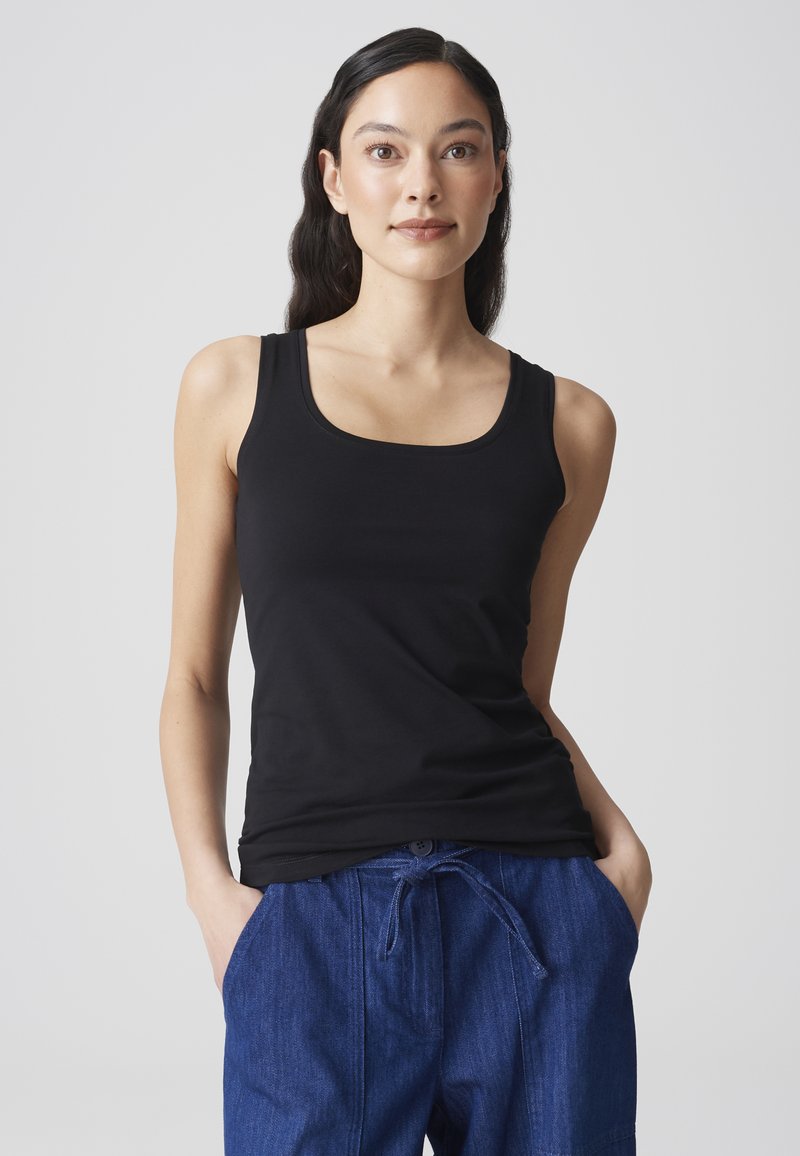 Jeune femme aux longs cheveux foncés portant un haut noir sans manches et un pantalon bleu avec cordon, sur un fond clair uni.