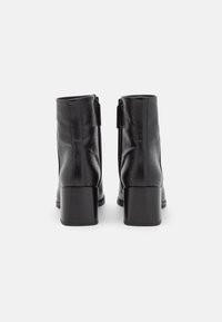 Botas de tobillo de imitación de piel negra con una superficie texturizada, tacón medianamente cuadrado y cierre de cremallera en la parte posterior. Acabado liso con patrones sutiles.