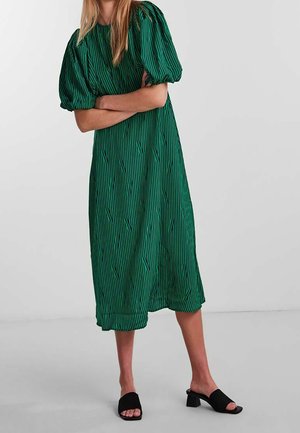 Vrouw draagt een groene midi-jurk met pofkorte mouwen en zwarte open-toe blokhak sandalen, staat met gekruiste armen.