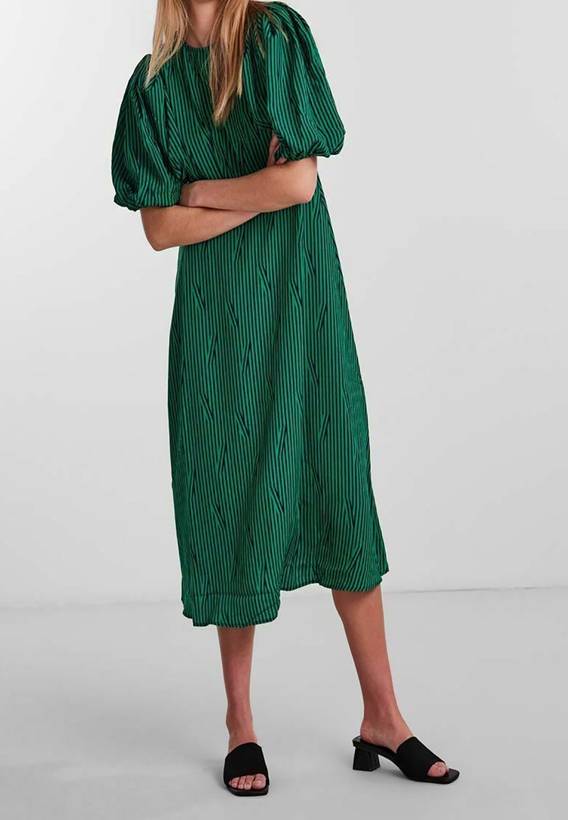 Femme portant une robe midi verte avec des manches courtes bouffantes et des sandales à talons blocs noires à bouts ouverts, debout les bras croisés.