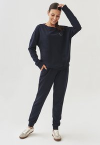 Sada tmavě modré mikiny a kalhot, která se vyznačuje uvolněným designem, měkkou texturou a žebrovanými manžetami. Boty jsou světle zbarvené s hnědými akcenty.