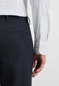 Vit skjorta med blå mönster, tillsammans med mörkblå byxor som har en bakficka med knappdetalj. Den figurnära designen framhäver formen.