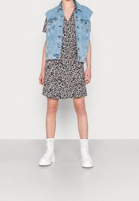 Giacca di denim senza maniche su un vestito nero a fiori con fiori bianchi. Il modello indossa stivaletti bianchi alla caviglia con lacci e suola spessa.