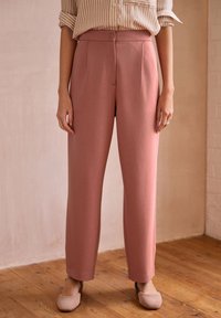 Rosa maßgeschneiderte Hose mit glatter Textur, hohem Bund, Falten und seitlichen Schlitztaschen, kombiniert mit hellen Schuhen.