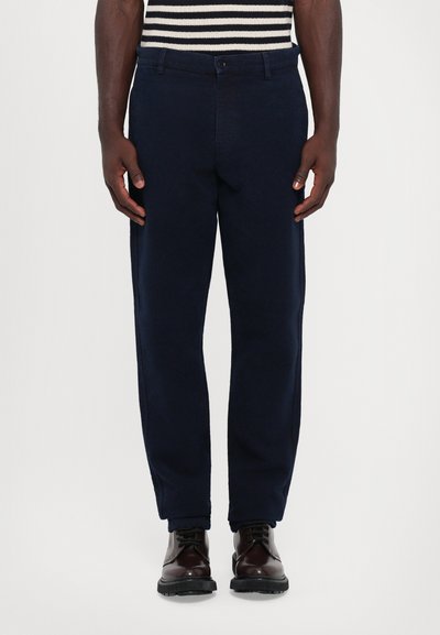 Pantalon bleu marine avec une texture lisse, design à jambes droites et poches latérales. Associé à des chaussures en cuir marron avec des semelles noires.