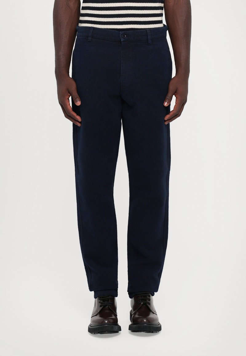 Marineblaue Hosen mit glatter Textur, geradem Bein-Design und seitlichen Taschen. Kombiniert mit braunen Lederschuhen mit schwarzen Sohlen.