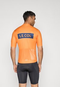 Oranžový cyklistický dres s krátkým rukávem, se znakem "LE COL" v námořnické modři, síťovanou zadní částí a kapsami na uložení. Nosený s černými šortkami.