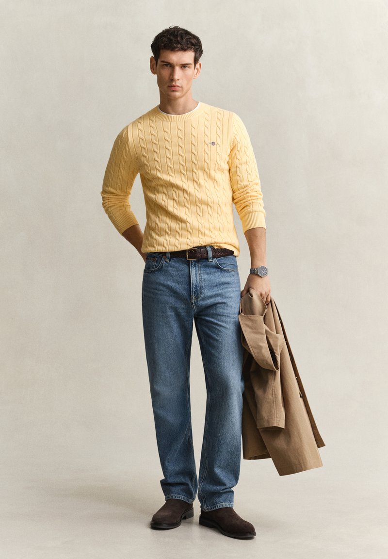 Homme portant un pull jaune à torsades et un jean bleu tient un manteau beige, debout sur un fond neutre de studio avec une expression sérieuse.