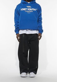 Felpa blu con la scritta "LOST YOUTH", tasca a marsupio, sovrapposta a una maglietta bianca. Pantaloni neri a gamba larga con un dettaglio di cinturino nero. Sneakers bianche.