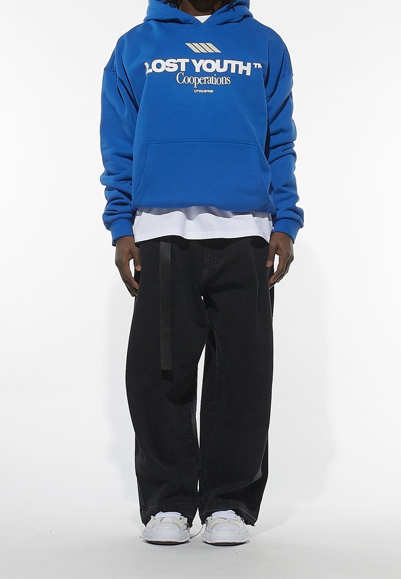 Felpa blu con la scritta "LOST YOUTH", tasca a marsupio, sovrapposta a una maglietta bianca. Pantaloni neri a gamba larga con un dettaglio di cinturino nero. Sneakers bianche.