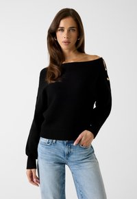 Zwarte gebreide trui met open schouders, lange mouwen en een geribbelde zoom; voorzien van goudkleurige hardware accenten bij de schouders. Gecombineerd met lichtblauwe jeans.