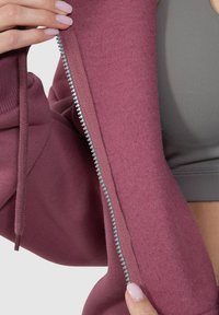 Felpa capospalla bordò con zip, caratterizzata da un interno morbido e testurizzato; cuciture a contrasto e uno strato base grigio visibile sotto.