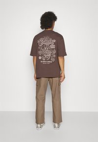 Osoba s kudrnatými vlasy stojí čelem ke zdi, na sobě má hnědé tričko s potiskem, béžové cargo kalhoty a tenisky s zebra pruhy.