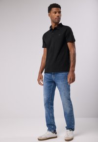 Mustat lyhythihaiset poolopaidat, joissa on kaulus ja logo, yhdistettynä vaaleansinisiin farkkuihin ja valkoisiin lenkkareihin, joissa on beige yksityiskohta.