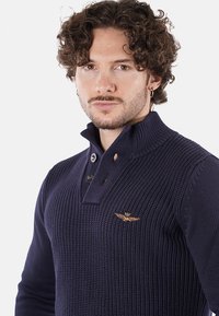 Maglione lavorato a maglia blu navy con colletto alto, dotato di tre bottoni e un logo ricamato in oro sul petto. Tessuto testurizzato e design aderente.