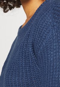 Pull en tricot bleu avec une finition texturée et un col rond, présentant un tissu doux et épais avec des détails de couture visibles.