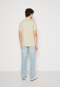 Μπεζ πόλο μπλούζα με κοντά μανίκια, λεία υφή, συνδυασμένη με ανοιχτό μπλε τζιν και λευκά αθλητικά παπούτσια, προσανατολισμένη από πίσω.