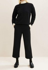 Personne portant un pantalon noir à jambes larges, un haut noir à manches longues par-dessus un col roulé blanc, des chaussettes noires et des mocassins noirs, debout les mains dans les poches.