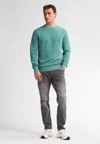 Turquoise gebreide trui met een ribbelstructuur, ronde hals, gecombineerd met grijze jeans en witte sneakers, staande op een neutrale achtergrond.