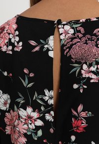Top negro con flores que presenta flores rosas y blancas, una abertura en la parte posterior con forma de llave y un cierre de botón en el cuello. Textura de tela suave.