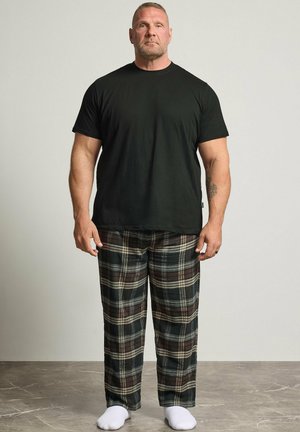 Homme debout portant un t-shirt noir, un pantalon à carreaux marron et noir, et des chaussettes blanches sur un fond clair et uni.