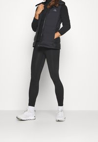 Černá bezrukávová péřová vesta s vysokým límcem, kombinovaná s černými legínami a bílými sportovními botami. Na vestě se nachází logo Adidas.