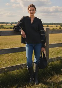 Schwarzes Langarmoberteil mit V-Ausschnitt und gepufften Ärmeln, kombiniert mit blauen Jeans und schwarzen kniehohen Stiefeln. Das Model hält eine schwarze Tasche.