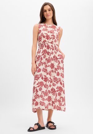 Femme portant une robe midi crème sans manches avec un imprimé floral rouge, debout, une main dans la poche et des sandales noires sur fond blanc.