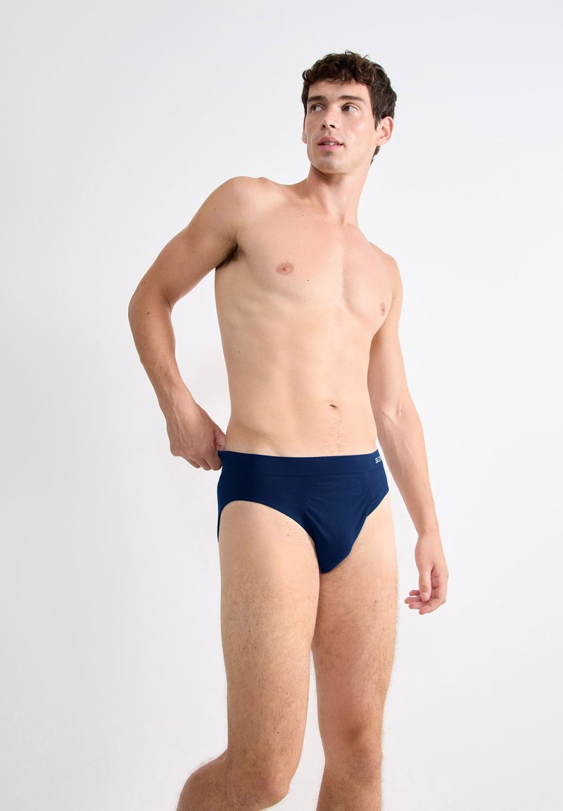 Oberkörperfreier Mann, der seine marineblauen Unterhosen anpasst, steht vor einem schlichten weißen Hintergrund und schaut nach links.